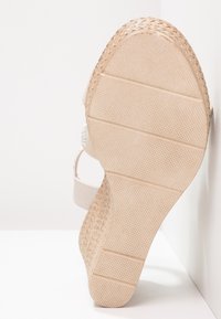 Sandale compensée haute avec une semelle beige texturée, un talon en jute tissé, et des brides en tissu clair ornées d'un accent métallique décoratif.