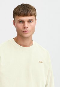 Cream-colored sweatshirt with a round neck and ribbed cuffs, featuring a small gold logo on the chest. Smooth, soft fabric texture.