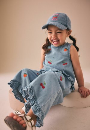 Young girl in blue denim outfit with fruit embroidery and a blue cap, laughing while sitting on a round beige stool.