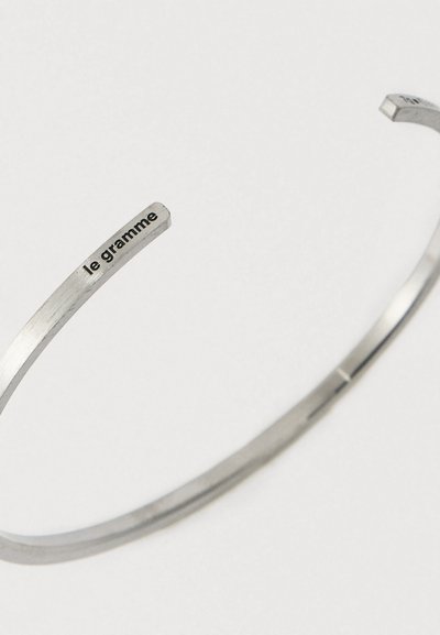 Sidabrinė atvira apyrankė su įspaudu „le gramme“ viename gale, pristatyta ant vienodo šviesaus fono.