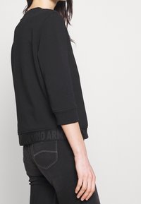 Černá mikina s tříčtvrtečními rukávy, žebrovanými lemy a texturovanou páskou s logem na lemu. Měkká látka a volný střih.