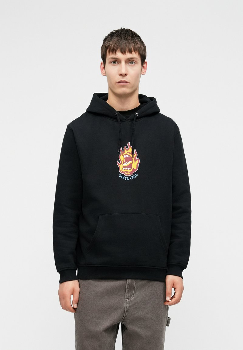 Μαύρη φούτερ με κουκούλα με μια μεγάλη, πολύχρωμη φλόγα και γραφικό στόμα στο κέντρο. Διαθέτει μπροστινή τσέπη και κουκούλα με κορδόνι. Μαλακή υφή.