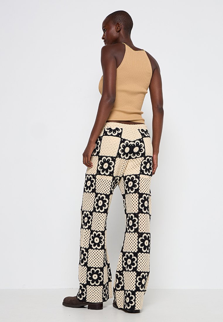 Beige gerippter Tanktop kombiniert mit weit geschnittenen Hosen im Häkelstil, die schwarze Blumenmuster auf cremefarbenem Hintergrund zeigen. Chunky braune Stiefel.