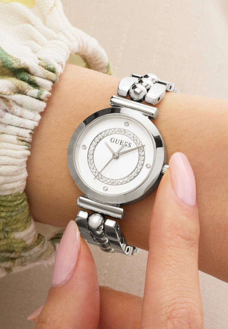 Ασημένιο ρολόι χειρός Guess με κρυσταλλικές διακοσμήσεις στη μεταλλική μπάντα, φορεμένο στον αριστερό καρπό, δάχτυλο που ρυθμίζει την κορώνα του ρολογιού, εμφανές μανίκι σε μπεζ-πράσινο χρώμα.