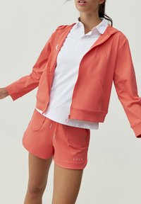 Koral hoodie med framzip och fickor, kombinerad med matchande shorts med dragsko och sidfickor, lager över en vit polo.