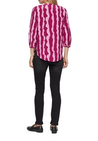 Vloeiende blouse met lange mouwen in een magenta en roze golvende streepjespatroon; ronde halslijn en losse pasvorm; gecombineerd met zwarte pantalons en platte schoenen.