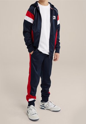 Granatowa bluza z zippem, ozdobiona czerwono-białymi paskami, w połączeniu z pasującymi granatowymi spodniami. Pod spodem znajduje się biała koszulka oraz sportowe buty.