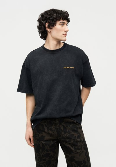 Νέος άνδρας με σγουρά μαλλιά που φοράει μαύρο T-shirt "Les Benjamins" και σκούρο παντελόνι με ανθικό σχέδιο, στέκεται με τα χέρια πίσω από την πλάτη, κοιτώντας προς τα δεξιά.