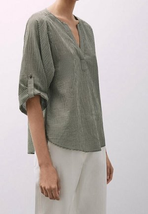 Femme portant un chemisier ample à carreaux avec manches trois-quarts retroussées et pantalon blanc sur un fond clair uni.