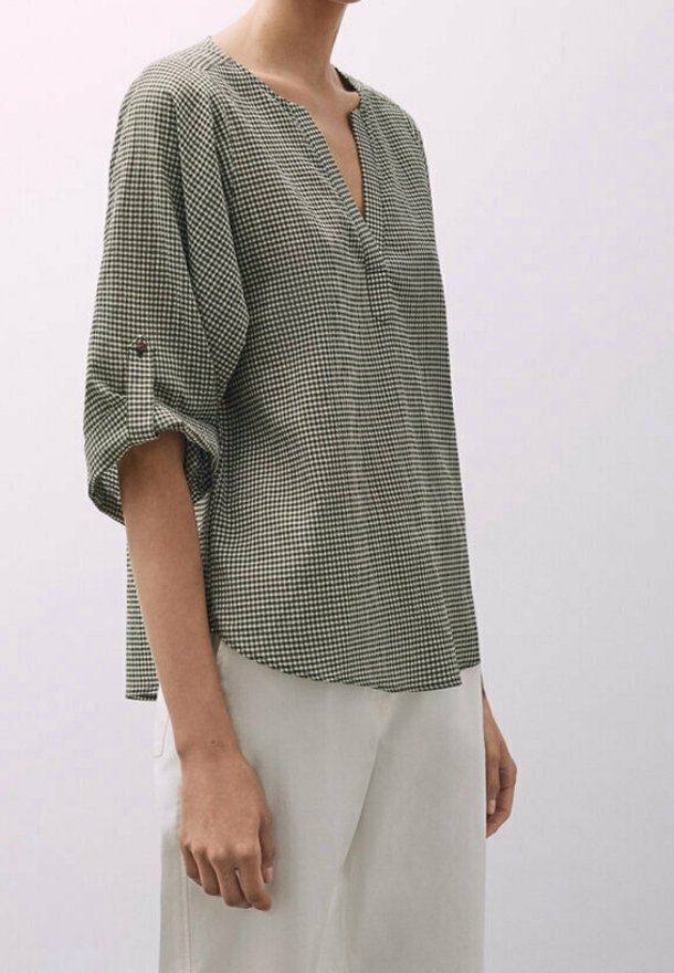 Femme portant un chemisier ample à carreaux avec manches trois-quarts retroussées et pantalon blanc sur un fond clair uni.