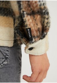 Marynarka z polaru w kratę w odcieniach brązu, kremu i czerni. Posiada oznaczoną mankiet, teksturowany materiał i zapięcie na guziki.