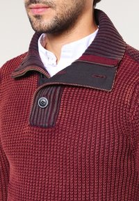Strickpullover in Burgunderrot mit strukturiertem Muster, ausgestattet mit einem dunklen Kragen, Knopfverschluss und Kontrastnähten.