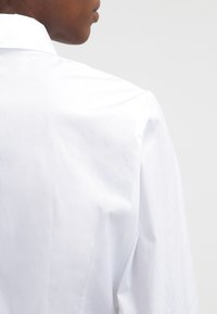 Camisa de algodón blanca con una textura suave, que presenta un cuello clásico y un ajuste entallado, mostrando la vista trasera y detalles de costura sutiles.