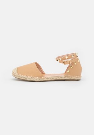 Espadrille plate beige à bout fermé, semelle en jute tressé, et bride de cheville ornée de clous pyramides dorés avec fermeture à boucle.