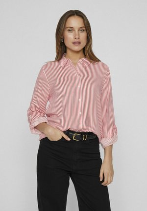 Blouse rayée rouge et blanche avec un col, un avant boutonné et des manches retroussées. Portée avec un jean noir et une ceinture noire.
