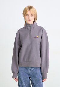 Persona rubia vistiendo un sudadera con cremallera gris de Carhartt y unos vaqueros azules holgados, de pie contra un fondo blanco liso.