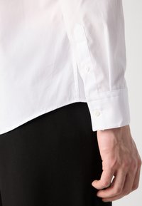 White collared shirt featuring buttoned cuffs, smooth texture, and a straight hem, paired with black trousers. Simple and classic design.