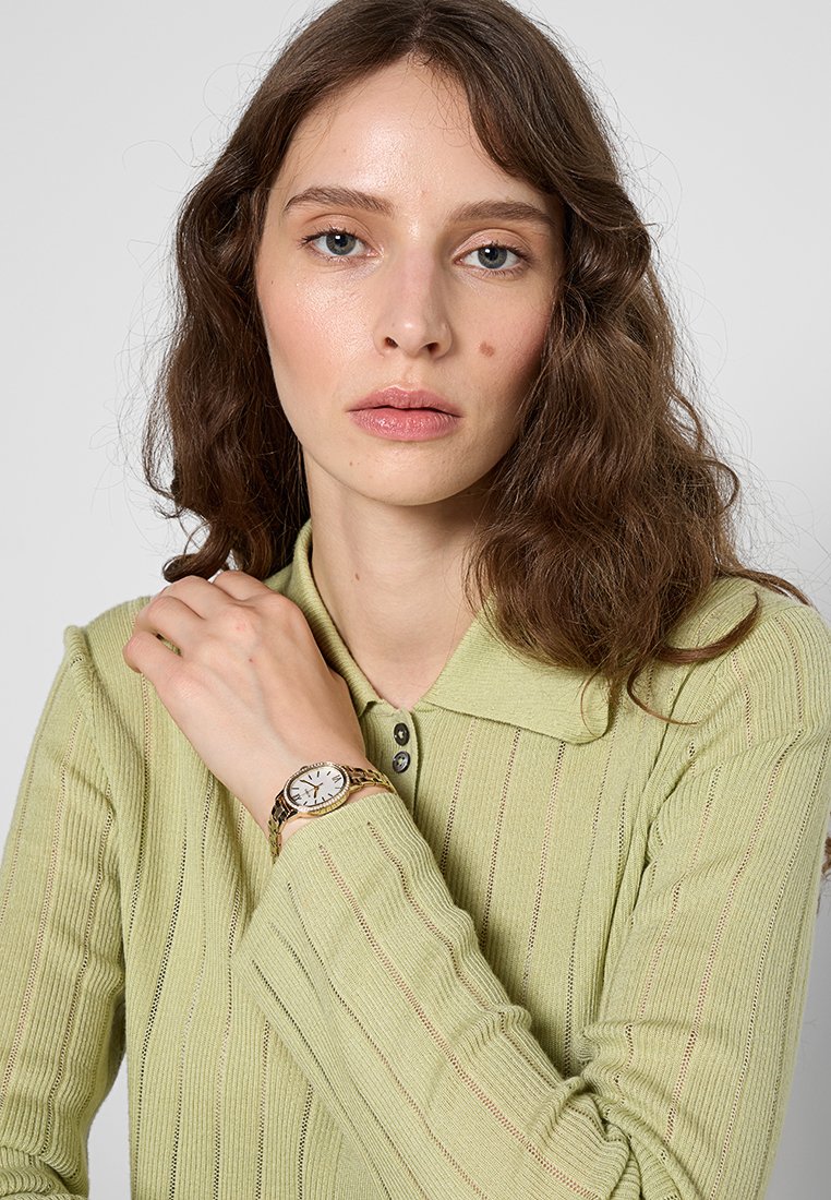 Un pull en maille côtelée vert clair avec un col, comportant des rayures verticales. Une montre-bracelet en ton or avec un cadran blanc est portée au poignet.