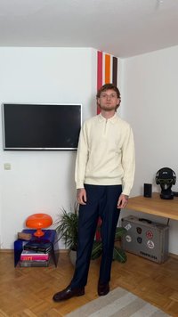 Ανοιχτό μπεζ ριγέ πουλόβερ με φερμουάρ και γιακά, ναυτικές παντελόνες και σκούρα καφέ παπούτσια. Εσωτερικός χώρος με πολύχρωμο τοίχο και έπιπλα.