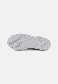 Semelle de chaussure blanche avec des crampons circulaires et un motif de bande de roulement en zigzag, vue du dessous sur un fond uni.