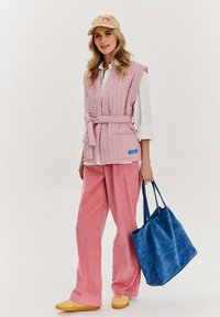 Różowa kamizelka pikowana z paskiem, biała koszula, różowe spodnie z wale woskowym, żółte buty wsuwane oraz niebieska torba na ramię. Na głowie czapka z logo.