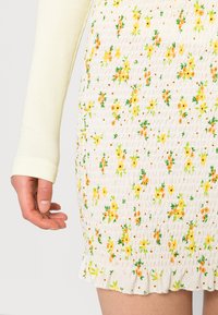 Vestido floral com um tecido branco franzido, apresentando flores amarelas e laranjas, folhas verdes e uma bainha com babados. Manga longa em creme com textura visível.