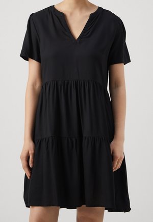 Kobieta ubrana w czarną sukienkę z krótkim rękawem, dekoltem w serek i tiulowym, marszczonym, warstwowym dołem, stojąca z luźno opuszczonymi ramionami.