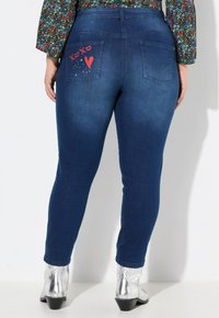 Jeans skinny de mezclilla azul oscuro con diseños de "XOXO" y corazones rojos en los bolsillos traseros, con un estilo de cinco bolsillos y una textura suave.