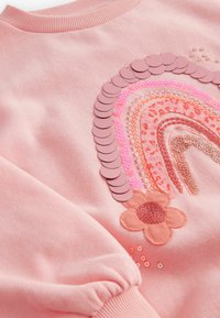 Tissu rose avec un arc-en-ciel brodé dans des nuances de rose et d'orange, décoré de paillettes et d'une fleur cousue en bas à droite.