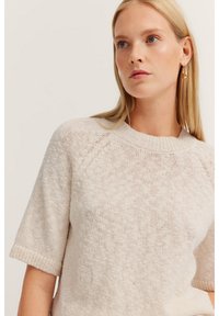 Kurzarmpullover mit rundem Halsausschnitt aus leichtem beigen Strickmaterial. Mit strukturiertem Muster und gerippten Bündchen für zusätzlichen Detailreichtum.