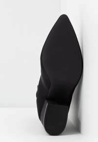 Chaussure noire à bout pointu avec une surface texturée, présentant un petit talon robuste et une semelle en caoutchouc lisse. Design minimaliste sans embellissements visibles.