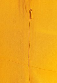 Gul texturerad tyg med en sydd söm och en liten gul dragkedjedetalj. Ytan verkar något skrynklig.