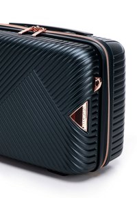 Valigia rigida verde scuro con texture rigata, zip oro rosa e accentuazione del logo triangolare. Forma rettangolare con finitura liscia.