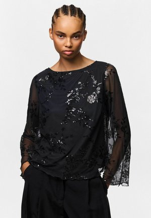 Jeune femme aux cheveux tressés porte un top noir transparent à manches longues avec des détails de sequins floraux et un pantalon noir sur fond blanc.