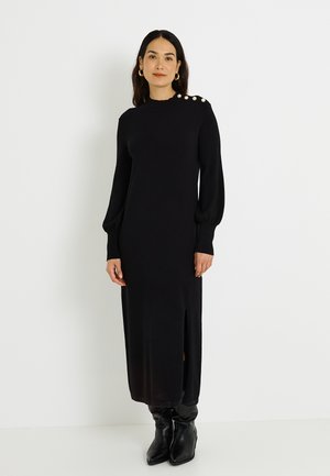 Robe noire en maille à manches longues avec boutons aux épaules, silhouette droite, fente latérale et poignets côtelés. Associée à des bottes noires montantes jusqu'au genou.