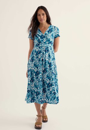 Femeie purtând o rochie midi albastră cu flori, cu mâneci scurte și sandale cu platformă bej, stând în fața unui fundal simplu, deschis la culoare.