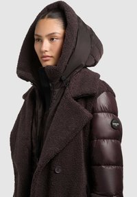 Tumepruun jakk, millel on pehme, tekstuuriga välispind, suur polsterdatud kapuuts ja läikiv varruka alumine osa. Sisaldab luku sulgemist ja logoga detaile.