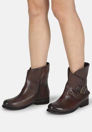Botas de cuero marrón hasta el tobillo con puntera redonda, cremallera lateral y una correa decorativa con hebilla. Textura suave, tacón bajo apilado.