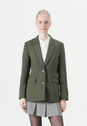 Blazer vert foncé avec revers crantés, design à un bouton, deux poches à l'avant et boutons argentés, associé à une chemise blanche et une jupe grise.