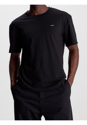 Mann trägt schlichtes schwarzes T-Shirt mit kurzen Ärmeln und kleinem Calvin Klein-Logo auf der Brust, die Hände teilweise in den Taschen der schwarzen Hose.