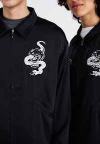 Črn zip-up jopič z vezenim dizajnom lobanje s kačo na prsih, gladka tkanina in stranski žepi.