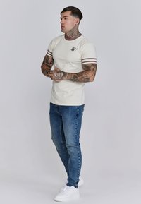 Crèmekleurig t-shirt met bruine gestreepte accenten op de mouwen, gecombineerd met blauwe jeans en witte sneakers, met een klein logo op de borst.