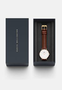 Hodinky Daniel Wellington v námorníckej krabici, s bielym ciferníkom, zlatými akcentami, hnedým koženým remienkom a minimalistickým dizajnom.