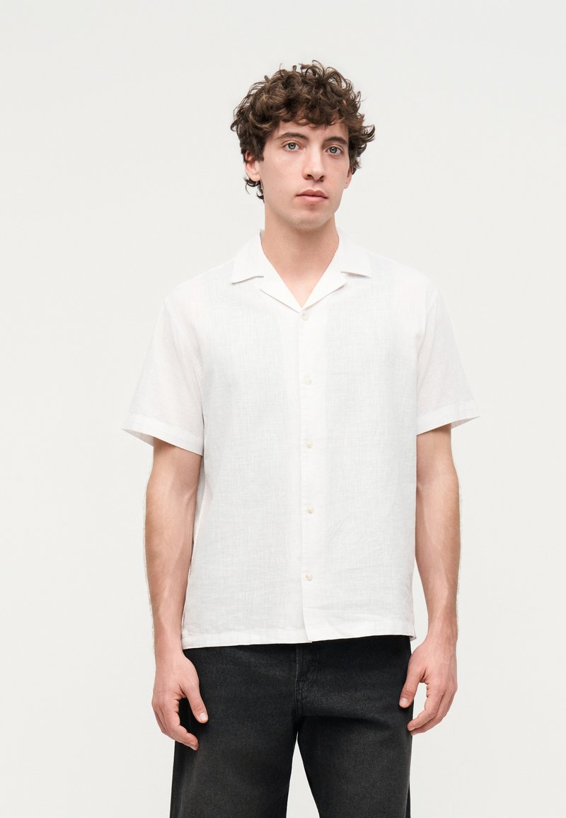 Νεαρός άνδρας με σγουρά μαλλιά που φοράει λευκό κοντομάνικο πουκάμισο με κουμπιά και μαύρο παντελόνι, στέκεται μπροστά σε ένα απλό φωτεινό φόντο.