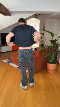 Zwart T-shirt met korte mouwen, grijze cargobroek met patroon en zijzakken, en zilveren sneakers; binnenomgeving met planten en meubels.