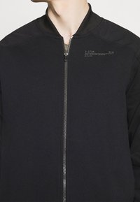Černá bomber bunda z hladké látky, s předním zipem, žebrovaným límcem a potiskem s logem na hrudi.