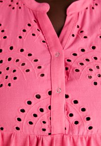 Vestido rosa com bordados em designs circulares recortados. Inclui uma frente com botões e uma barra com volantes. Textura suave.