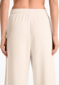 Beige, wide-leg pants with an elastic waistband. Smooth texture, no visible patterns or hardware, shown from the back with hand positioned on hip.