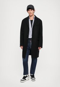 Black wool overcoat with three buttons, gray shirt underneath, blue denim jeans, and black and white sneakers, featuring a baseball cap.