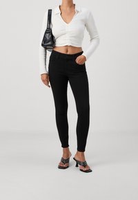 Jeans negros ajustados de cintura alta, combinados con una blusa blanca entallada de canalé con cuello y detalles fruncidos en la parte delantera. Llevados con mule negros de tacón.